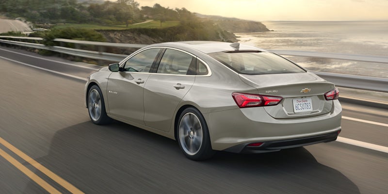 A Chevrolet Malibu driving along a scenic road surrounded by trees and greenery.