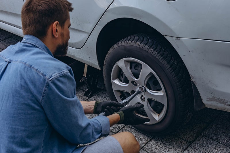 A man kneels beside a car, repairing a flat tire with tools in hand.