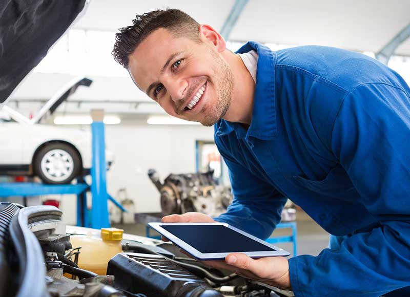 A man examines his car while holding a tablet in his hands, focused on the device's screen.